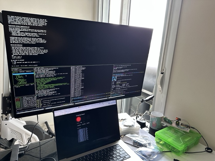 A wide shot of the working desk: vertical monitor showing a tmux split with code on the left and Nerves CLI on the right, a laptop below, oscilloscope and bench tools in the background, parts bin to the side.