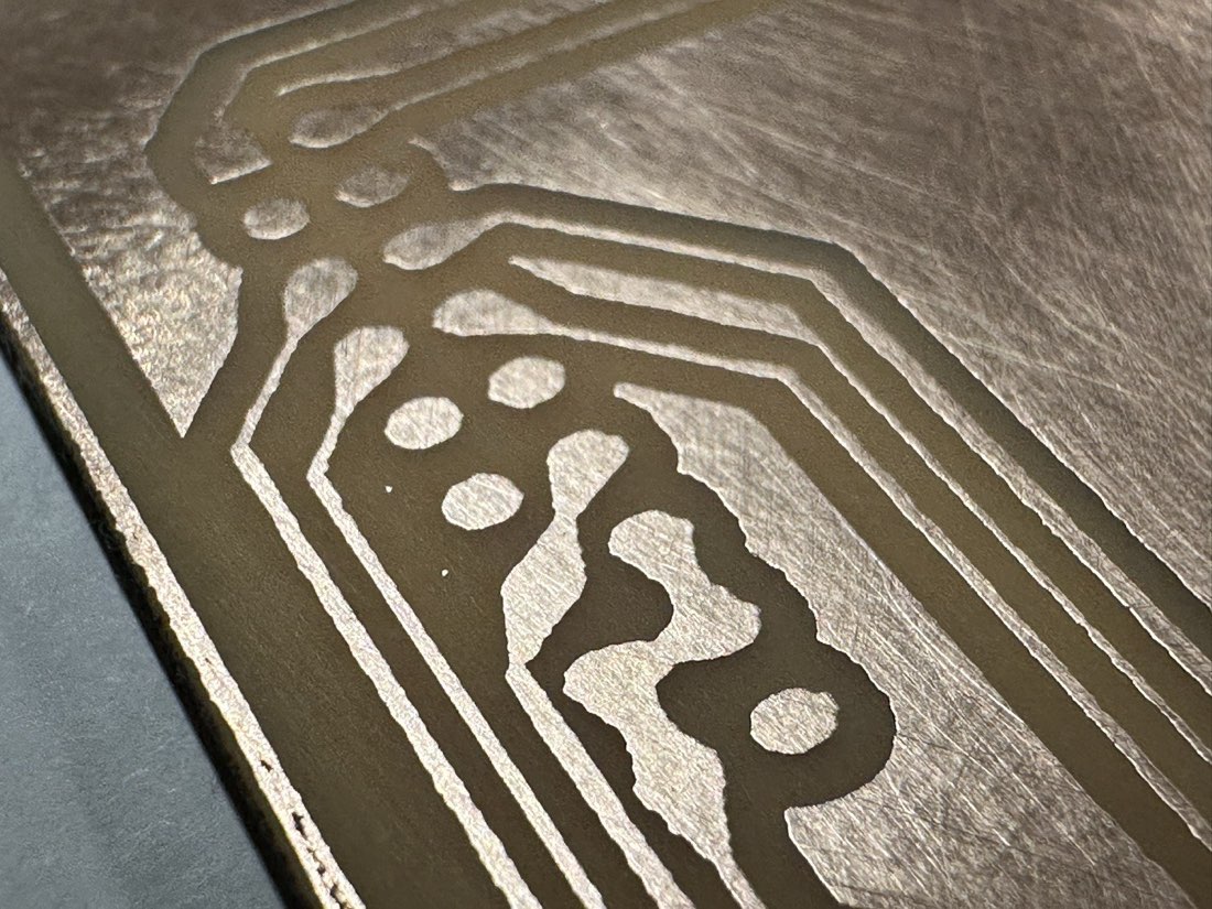Extreme close-up of etched copper traces with ragged, irregular edges and pitted copper around them, the resist coating still on top, showing the undercutting from a long etch in a weak bath.