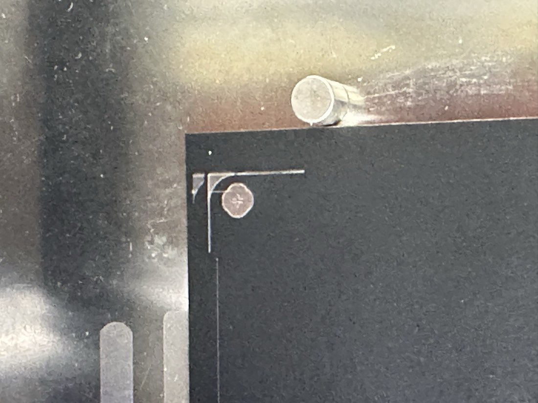 Macro view of a board corner showing a lasered cross-hair fiducial mark near the corner, a metal bed clamp post beside it, the board held on the printer bed.
