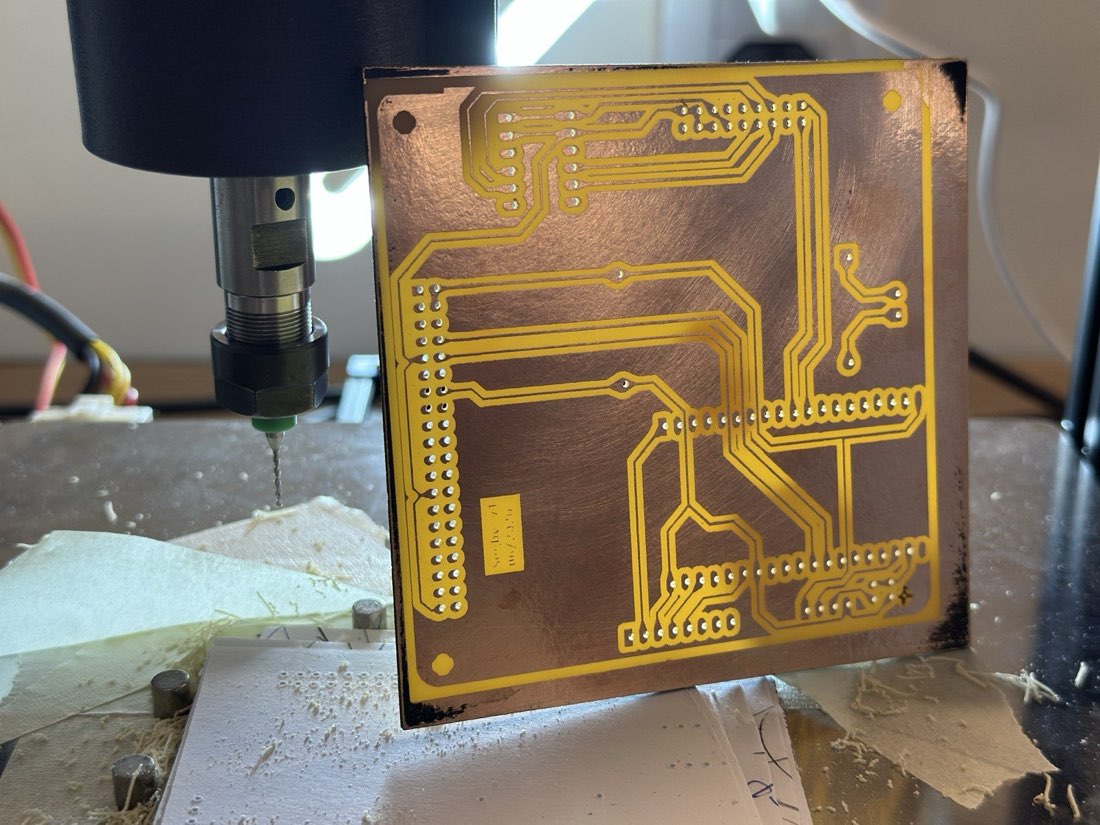 The finished etched-and-drilled segby_v1 board standing on the printer bed, backlit so the drilled holes glow, the 775 spindle and drill bit just behind it, gold-toned traces on copper with FR4 dust and bed magnets around it.