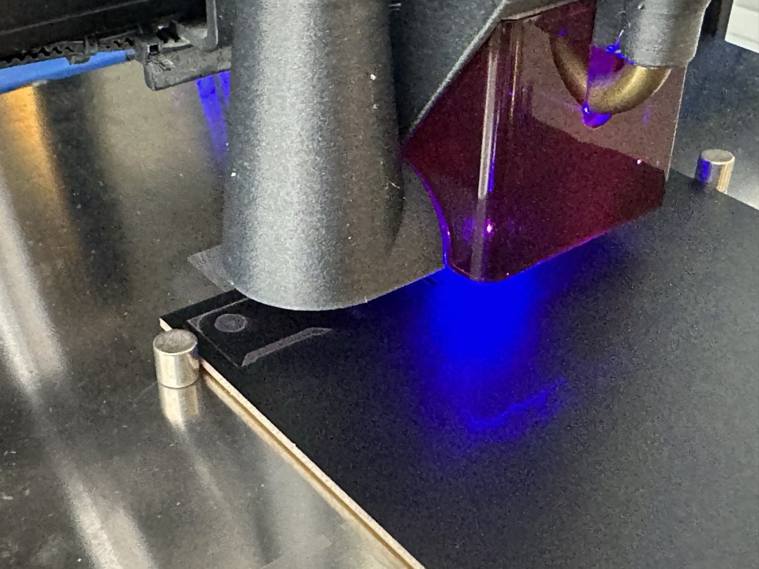 The diode laser firing a bright blue-violet beam down onto a black-painted copper board clamped on the printer bed, the printed laser shroud glowing red-violet around the beam.