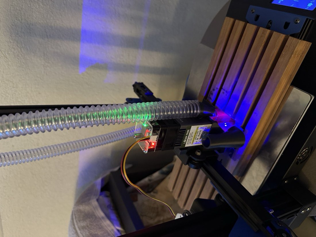 Close-up of the Two Trees Bluer print head with a blue diode laser module bolted to the carriage next to the hotend, ribbed exhaust ducting running up to it, small red and green status LEDs lit on the driver board, the bed lit blue underneath.