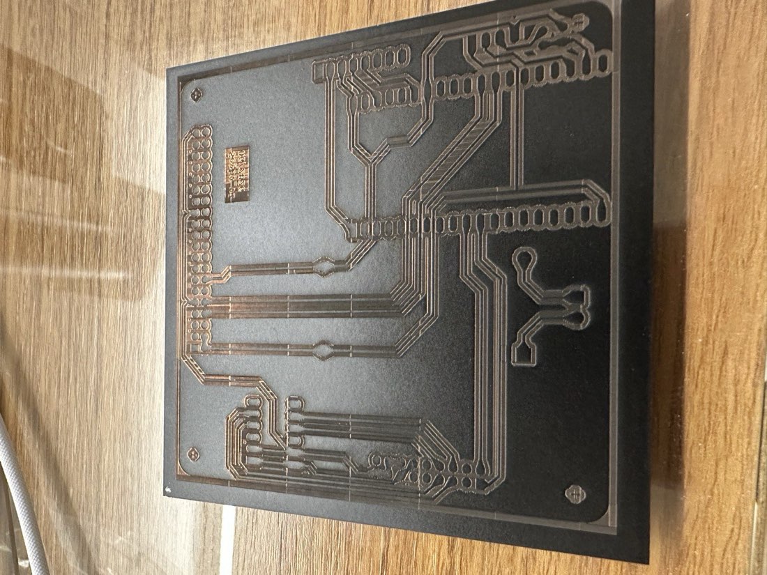 A black-painted board with the full PCB trace pattern ablated into it, bare copper traces visible as lighter lines against the remaining black paint, sitting on a wooden desk.