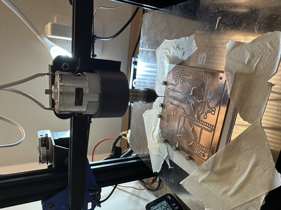 The 775-motor spindle mounted on the printer, drill bit plunging into an etched copper board surrounded by paper towel and a scatter of FR4 dust, the board held down by bed clamps.