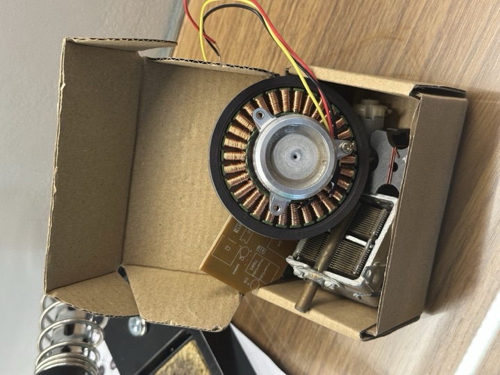 A 30-slot floppy-drive spindle motor with its windings exposed, sitting in a small cardboard box on a wooden desk, three soldered wires already coming out of the stator.