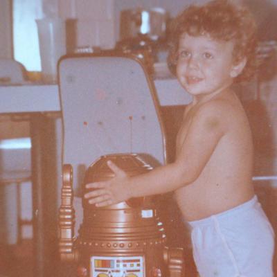 A small child with curly hair, smiling, hands on the lifted lid of a toy robot taller than they are. The robot is the original Ar-tur: brown plastic body, control panel on the chest, antenna up.